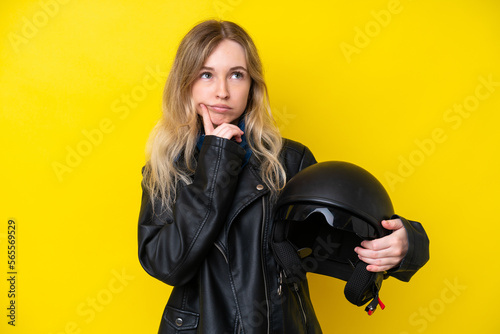 Blonde English young girl with a motorcycle helmet isolated on yellow background having doubts while looking up