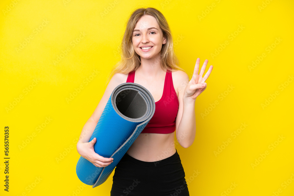 Young sport caucasian woman going to yoga classes while holding a mat isolated on yellow background happy and counting three with fingers