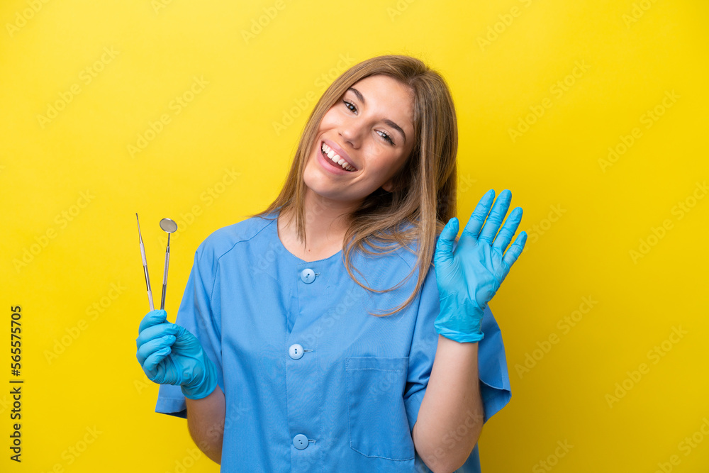 Dentist caucasian woman holding tools isolated on yellow background saluting with hand with happy expression Dentist caucasian woman holding tools isolated on yellow background saluting with hand with happy expression