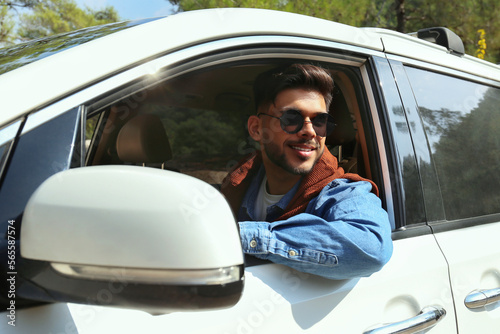 Young carefree man wearing a denim shirt looking out of the window of white car. Smiling guy enjoying his road trip. Close up, copy space, background. © Evrymmnt