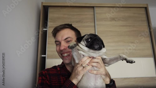 A young man in a red shirt communicates via video communication via the Internet at home. View from the laptop webcam. The man greets, shows his pug, smiles, laughs.