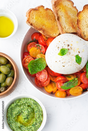Farmer Italian soft cheese mozzarella di bufala campana served with fresh basil and red and yellow cherry tomatoes, croutons, homemade pesto, olives. Healthy Italian summer food, white table, close-up