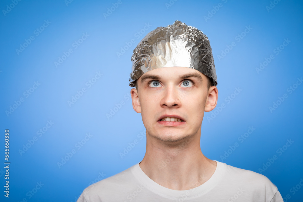 Fototapeta premium Paranoid young man wearing tin foil hat studio shot on blue background