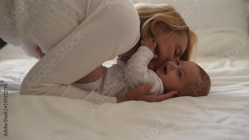 Smiling baby and mother in white clothes play on bed. Side view. mother kiss and laugh with baby. love and care in happy family. First year of life and knowledge of world around.