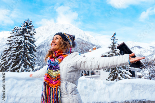 Overjoyed happy young woman in snow scenic place with chalet house and trees with mountains in background opening arms outstretching. Female people and winter holiday vacation travel leisure activity photo