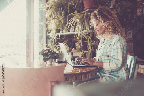 Side view of young woman sitting at the desk and writing on laptop. One businesswoman in modern smart working. Garden plants store shop. Female people using computer indoor to work and enjoy business © simona
