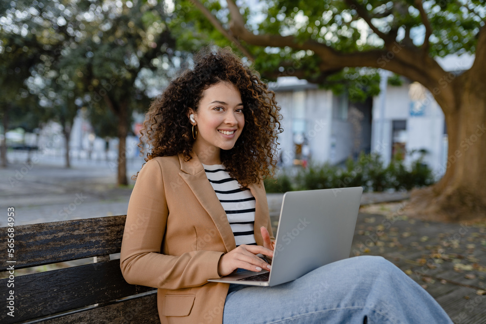 pretty curly woman sitting in city street in stylish jacket, working remote job on laptop pretty curly woman sitting in city street in stylish jacket, working remote job on laptop
