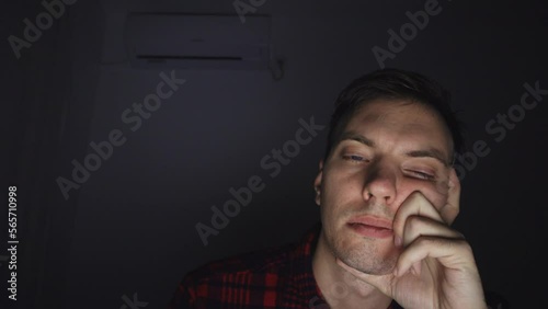Portrait of a young man falling asleep in front of a laptop camera during a night video call. The man is overworking, he is tired. The room is dark, the face illuminates the computer screen.