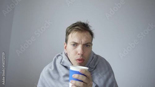 A sick person is sitting in front of a laptop, wrapped in a blanket, during a video call, a business conference, drinking hot tea, coughing. View from the laptop webcam.