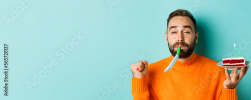 Close-up of funny adult man celebrating his birthday, holding bday cake with candle, blowing party wistle and rejoicing, standing over light blue background photo