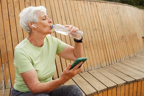 An elderly happy pensioner woman after a run sits on a wooden bench and drinks water. photo