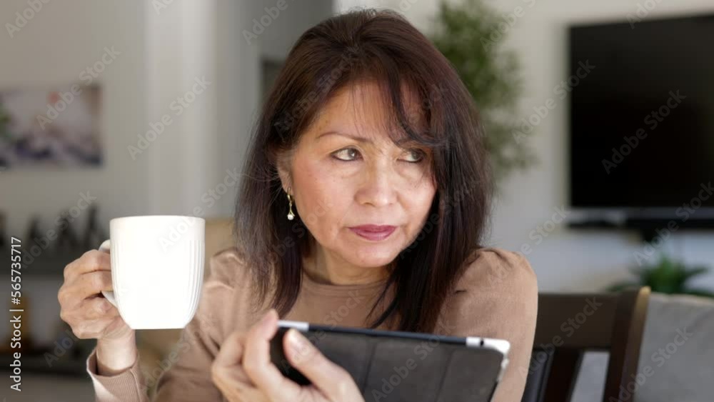 Close up attractive middle age woman relaxing drinking coffee reading mobile tablet looking sitting at table in casual living room of leisure home Close up attractive middle age woman relaxing drinking coffee reading mobile tablet looking sitting at table in casual living room of leisure home