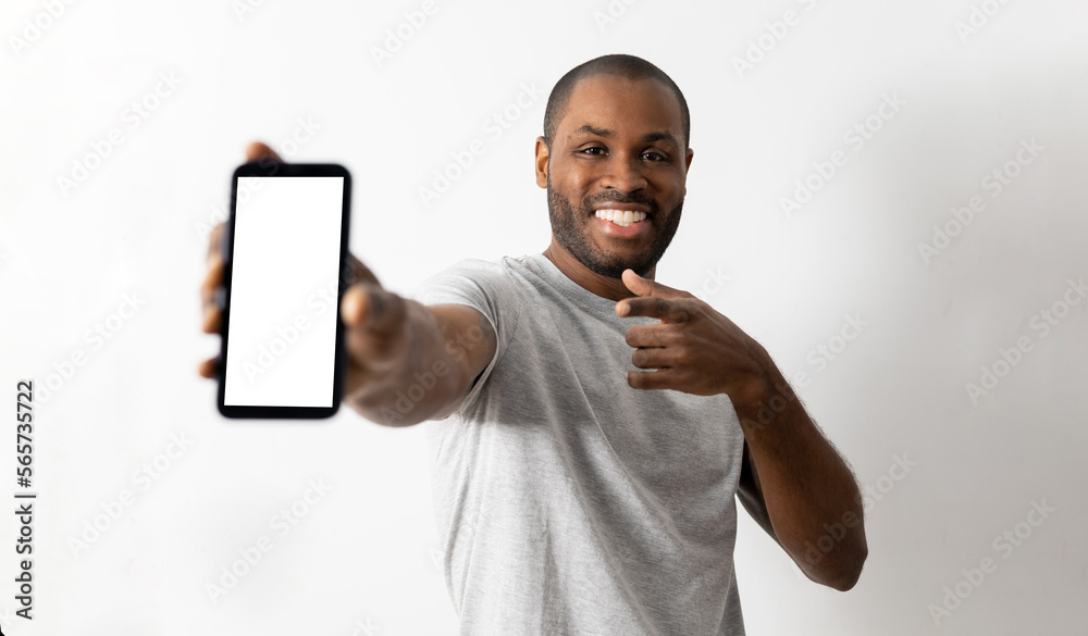 Satisfied smiling dark-skinned man showing the white screen mobile phone to the camera standing isolated in the white space of the studio. The man points to the phone with his finger. Advertising . Satisfied smiling dark-skinned man showing the white screen mobile phone to the camera standing isolated in the white space of the studio. The man points to the phone with his finger. Advertising .