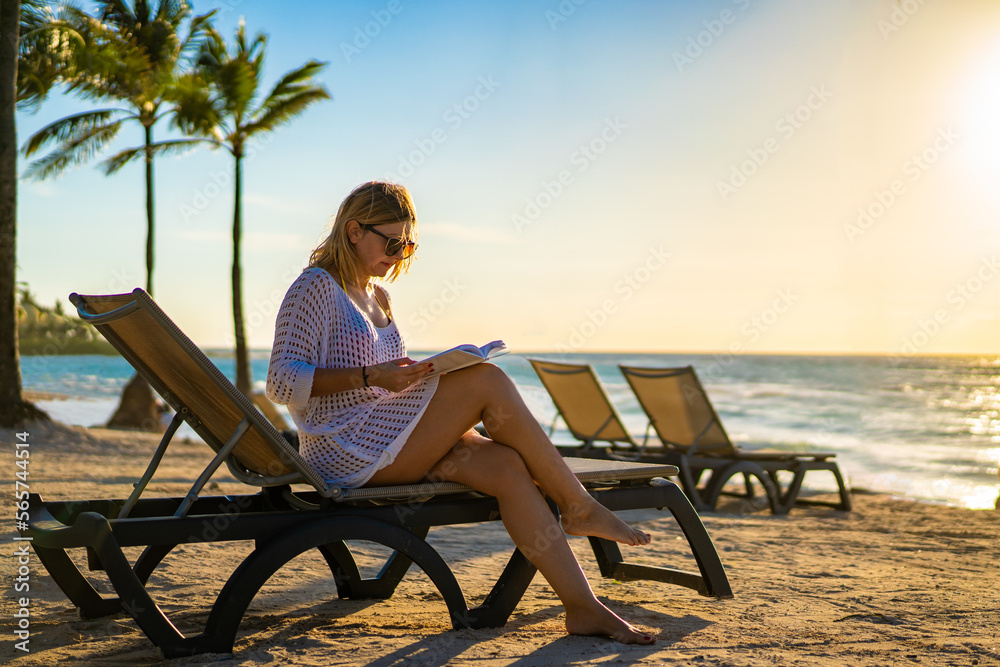Beach holiday - woman relaxing on beach reading book sitting on sunbed

