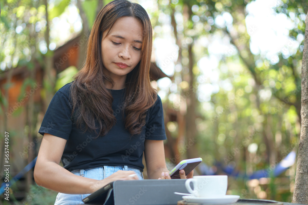 Fototapeta premium Asian women are happy to sit and work in a coffee shop. There are green trees surrounded by nature. Business planning is a good idea.