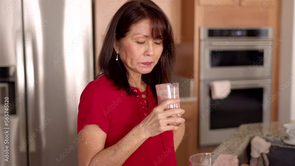 Attractive healthy mature woman standing drinking fresh fruit smoothie in kitchen of leisure home for morning breakfast Attractive healthy mature woman standing drinking fresh fruit smoothie in kitchen of leisure home for morning breakfast