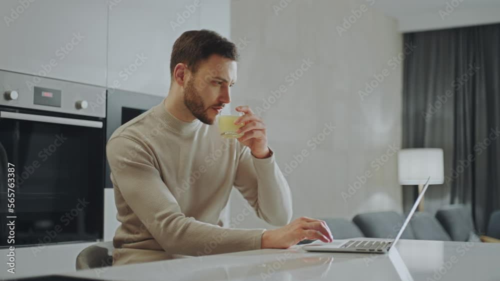 Bearded young man sitting at his desk and drinking juice while typing on laptop keyboard. Man working remotely
