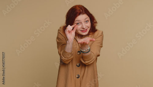 Need some more, please give me. Redhead woman showing a little bit gesture with sceptic smile, showing minimum sign, measuring small size. Young ginger girl isolated alone on beige studio background