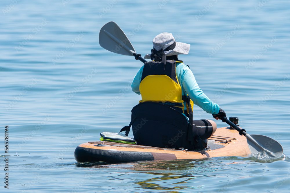 Naklejka premium Woman in life jacket and on SUP board exercising at sea water. Special equipment such as seating chare and wet suit on paddleboard. Training and sport on paddle SUP board at sunny summer day.