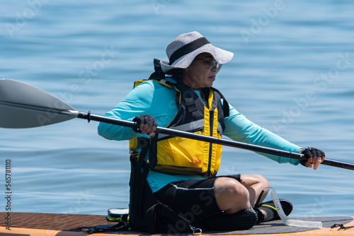 Woman in life jacket and on SUP board exercising at sea water. Special equipment such as seating chare and wet suit on paddleboard. Training and sport on paddle SUP board at sunny summer day.