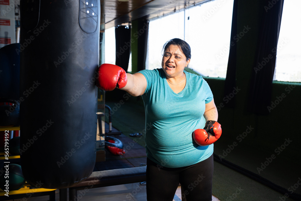 Angry overweight indian woman wearing boxing gloves punching bag in gym, Plus size sporty asian female workout to lose weight. Fitness and healthcare.