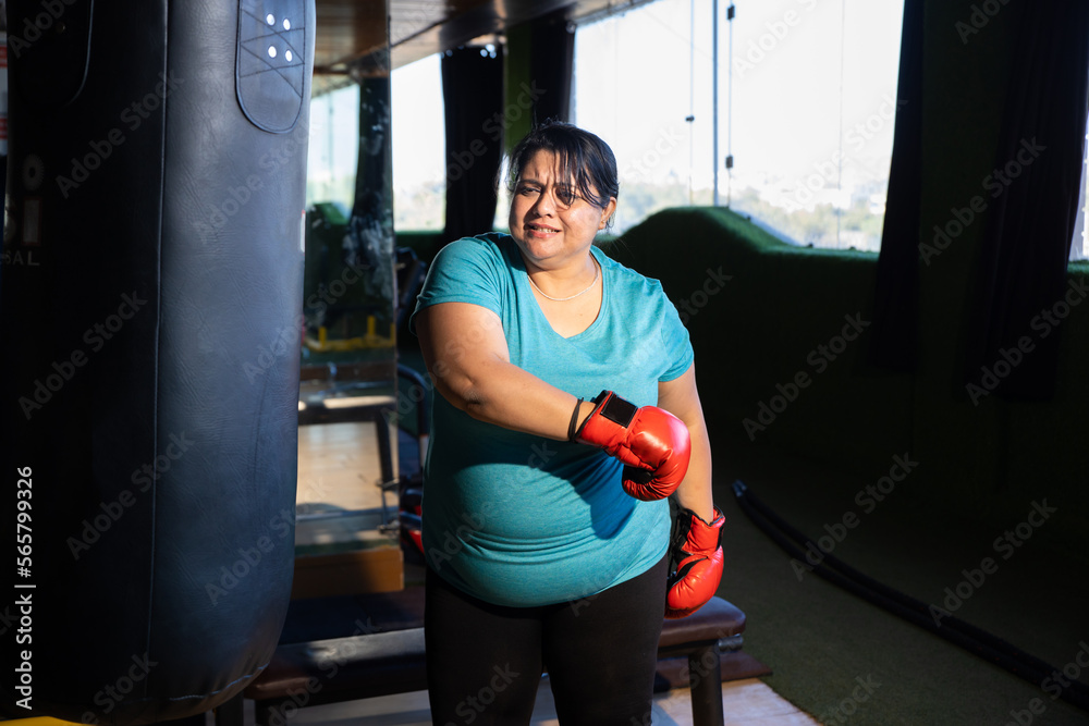 Frustrated Young overweight indian woman wearing boxing gloves punching bag in gym, Plus size sporty asian female workout to lose weight. Fitness and healthcare.