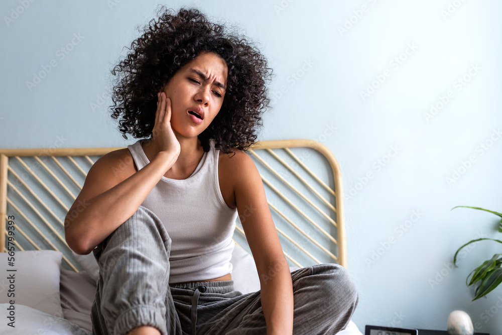 © Daniel - Young latina female with curly hair touching cheek in pain having toothache sitting on bed in cozy bedroom at home. © Daniel - Young latina female with curly hair touching cheek in pain having toothache sitting on bed in cozy bedroom at home.