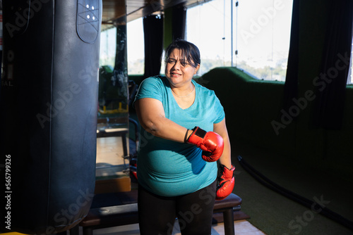 Frustrated Young overweight indian woman wearing boxing gloves punching bag in gym, Plus size sporty asian female workout to lose weight. Fitness and healthcare.