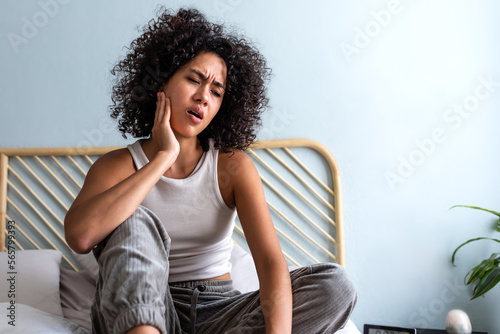Young latina female with curly hair touching cheek in pain having toothache sitting on bed in cozy bedroom at home.