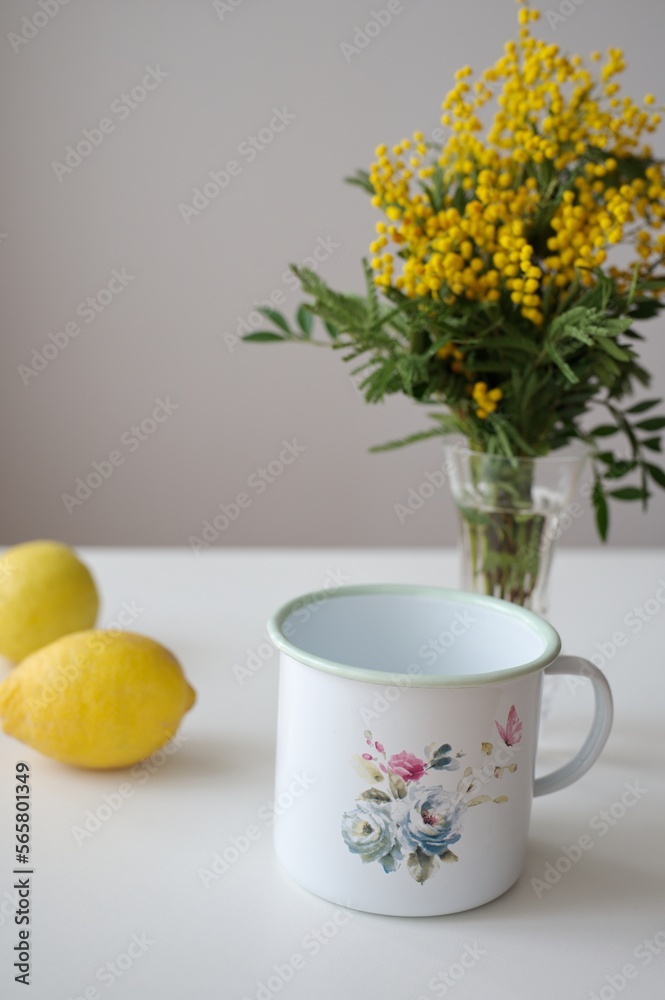 Enameled painted pot, lemon and yellow mimosa flowers in a glass vase on white table