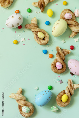 Happy Easter Holidays. Colors Easter eggs with Easter rabbit-shaped buns puff pastry with cinnamon on pastel green background. View from above. Copy space.