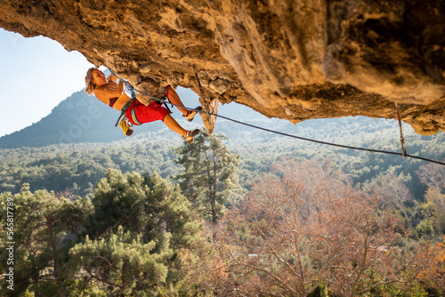 girl rock climber climbs a rock against the backdrop of a forest and blue mountains. climber goes through a difficult route using equipment.