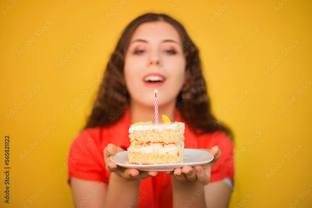 Selective focus on a piece of cake with a candle on a white plate in the hands of a girl in a red shirt isolated on a yellow background. Birthday celebration concept. Selective focus on a piece of cake with a candle on a white plate in the hands of a girl in a red shirt isolated on a yellow background. Birthday celebration concept.