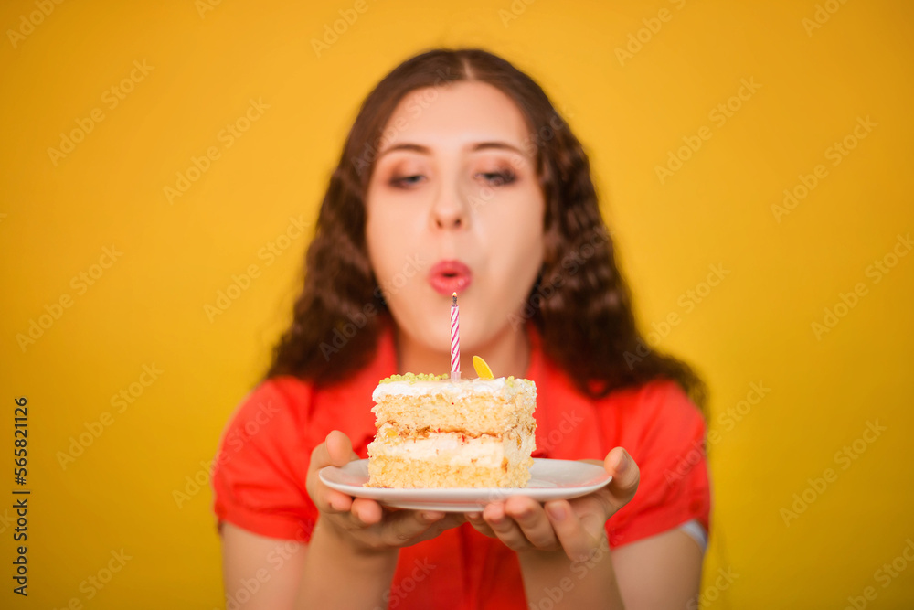 Selective focus on a piece of cake with a candle on a white plate in the hands of a girl in a red shirt isolated on a yellow background. Birthday celebration concept.