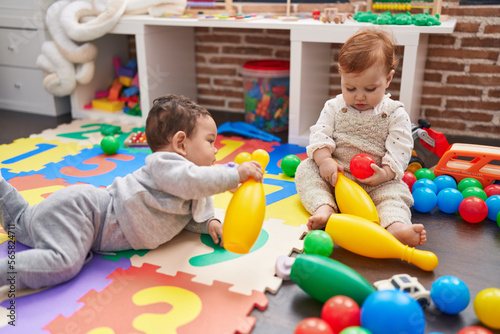 Two adorable babies playing with balls and bowling pin sitting on floor at kindergarten