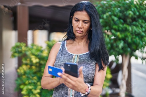 Middle age hispanic woman using smartphone and credit card with serious expression at street