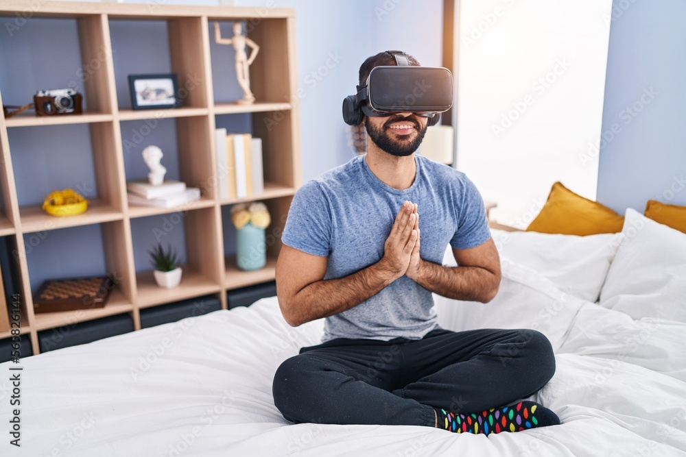 Young hispanic man doing yoga exercise using virtual reality glasses at bedroom
