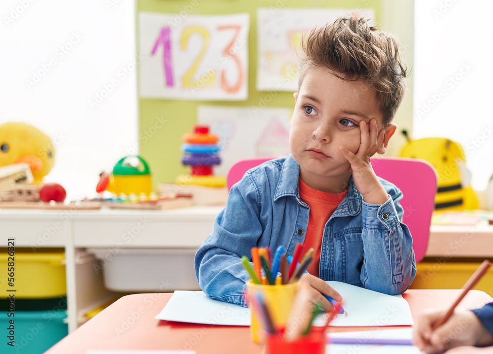 Adorable hispanic toddler student sitting on table drawing on paper with doubt expression at kindergarten Adorable hispanic toddler student sitting on table drawing on paper with doubt expression at kindergarten