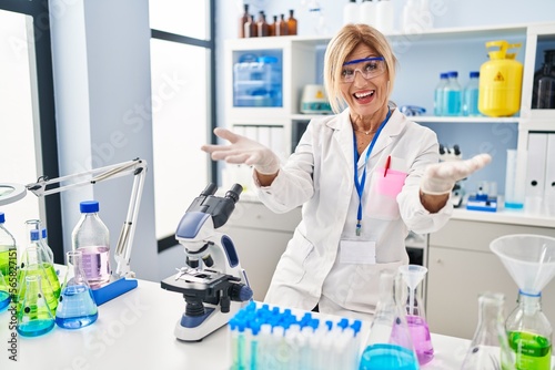 Middle age blonde woman working at scientist laboratory smiling cheerful offering hands giving assistance and acceptance.