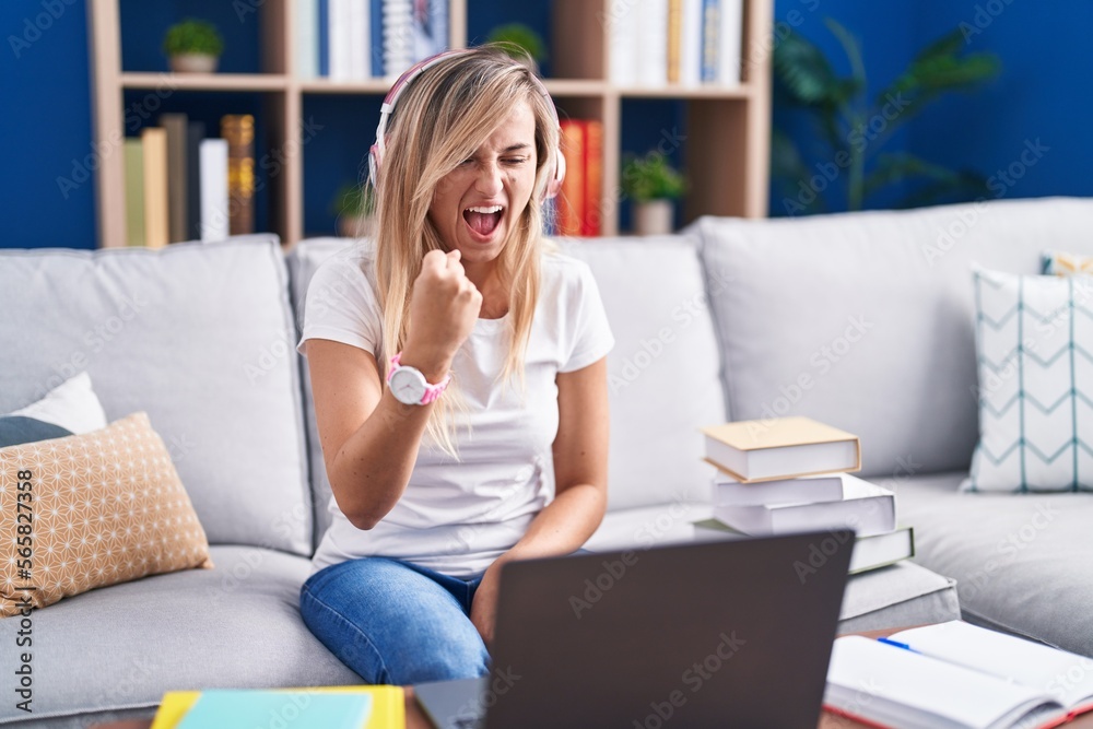Young blonde woman studying using computer laptop at home angry and mad raising fist frustrated and furious while shouting with anger. rage and aggressive concept. Young blonde woman studying using computer laptop at home angry and mad raising fist frustrated and furious while shouting with anger. rage and aggressive concept.