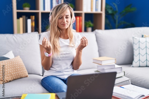 Young blonde woman studying using computer laptop at home doing money gesture with hands, asking for salary payment, millionaire business
