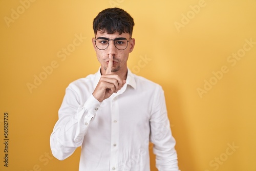 Young hispanic man standing over yellow background asking to be quiet with finger on lips. silence and secret concept.