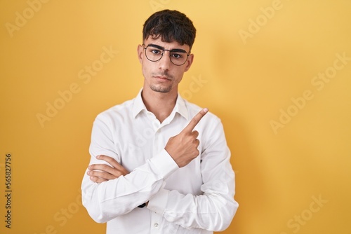 Young hispanic man standing over yellow background pointing with hand finger to the side showing advertisement, serious and calm face © Krakenimages.com