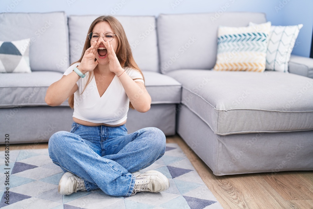 Young caucasian woman sitting on the floor at the living room shouting angry out loud with hands over mouth Young caucasian woman sitting on the floor at the living room shouting angry out loud with hands over mouth