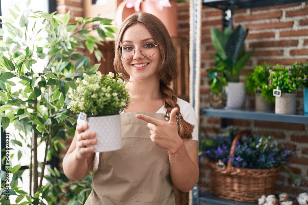 Young caucasian woman working at florist shop holding plant smiling happy pointing with hand and finger Young caucasian woman working at florist shop holding plant smiling happy pointing with hand and finger