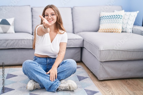 Young caucasian woman sitting on the floor at the living room smiling friendly offering handshake as greeting and welcoming. successful business.
