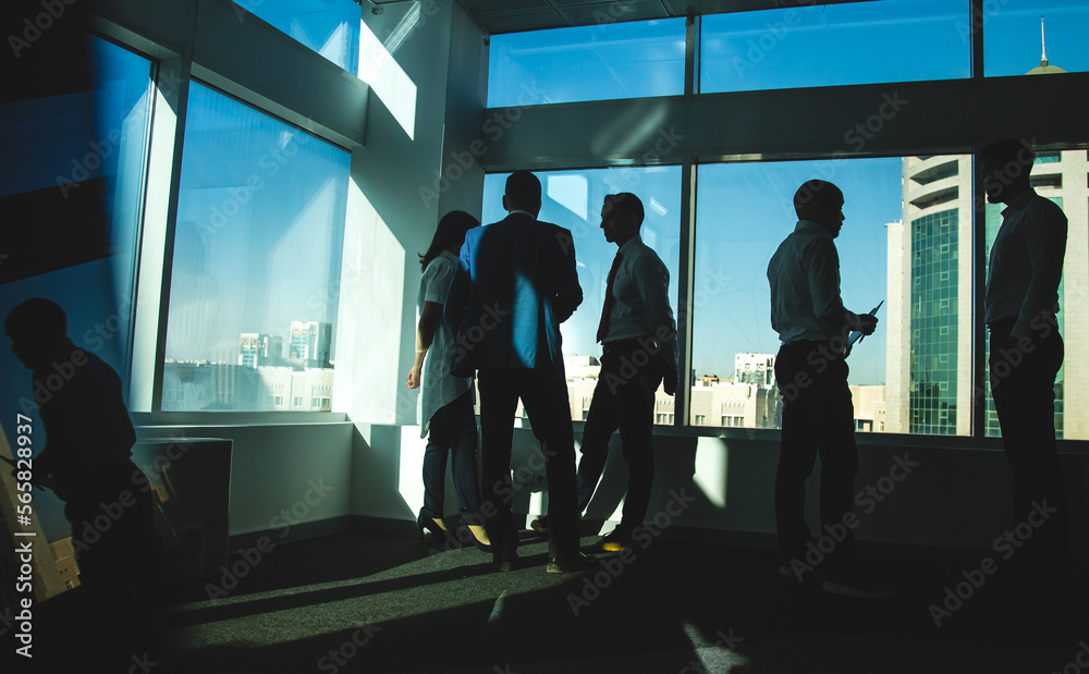 Silhouettes of people standing near a panoramic window in a modern office. Team of young professional business people working and chatting together in a meeting.
