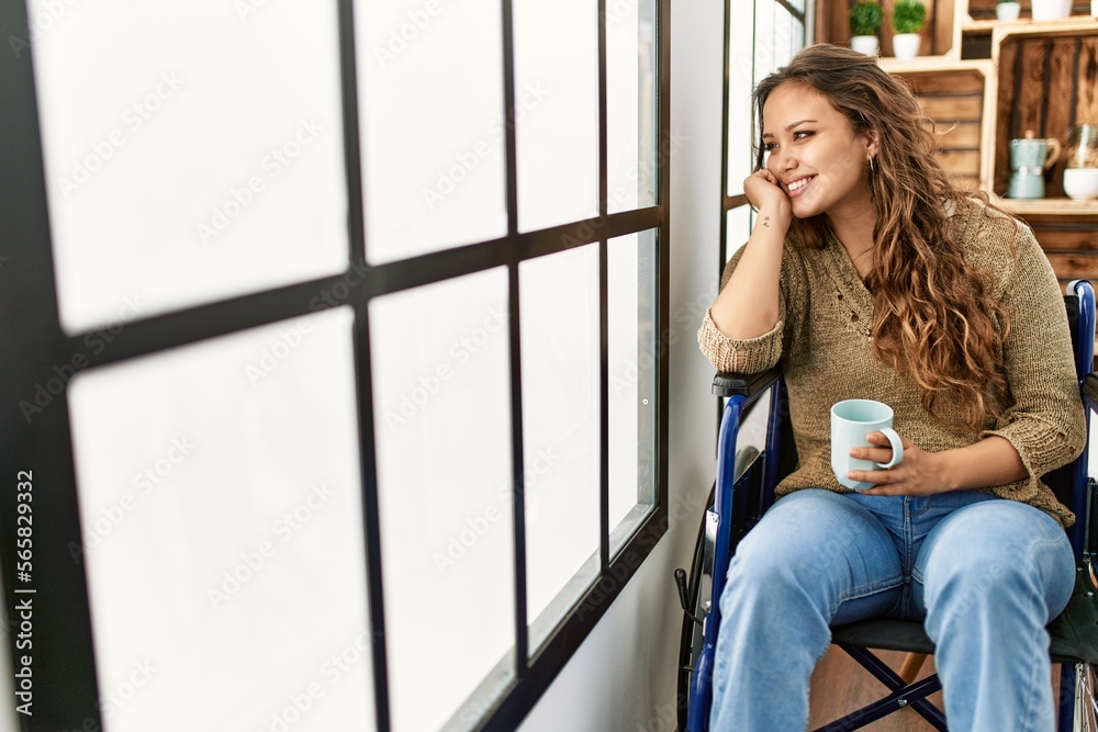Young beautiful hispanic woman drinking coffee sitting on wheelchair at home