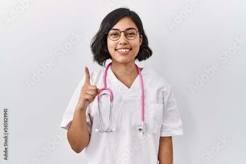 Young hispanic doctor woman wearing stethoscope over isolated background showing and pointing up with finger number one while smiling confident and happy.