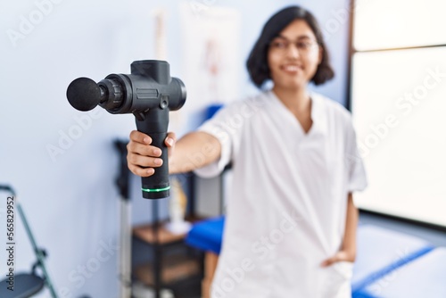 Young latin woman wearing physiotherapist uniform holding percussion pistol at physiotherapy clinic